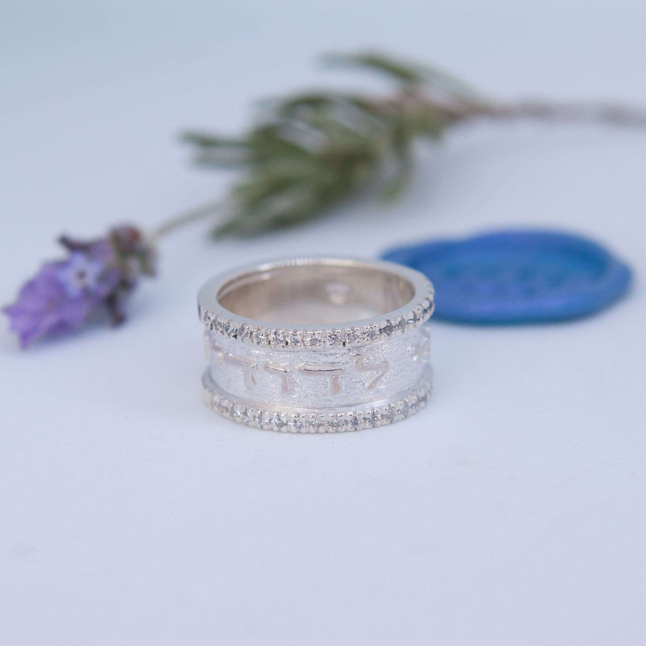The image shows a silver ring with a textured, matte finish and tiny stones set along its edges. The ring has raised Hebrew lettering around its band.